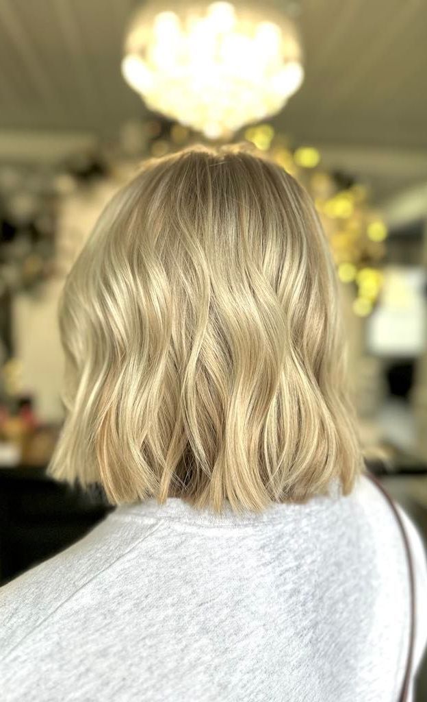 Blonde bob hairstyle with soft waves, viewed from the back.  The person is wearing a gray sweater.  A blurred light fixture hangs above.