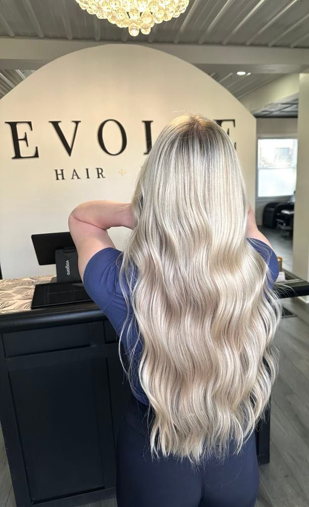 Woman with long wavy blonde hair at the Evolve Hair salon, arms raised behind her head, dark shirt, light setting.