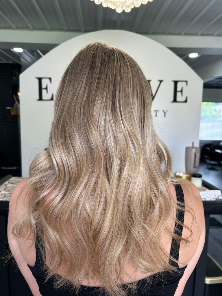 Woman with wavy, highlighted blonde hair in a salon. The salon has a white arch with the name 