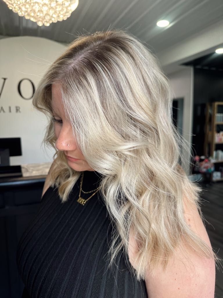 Woman with blonde wavy hair and dark roots in a salon, wearing a black top, looking down.