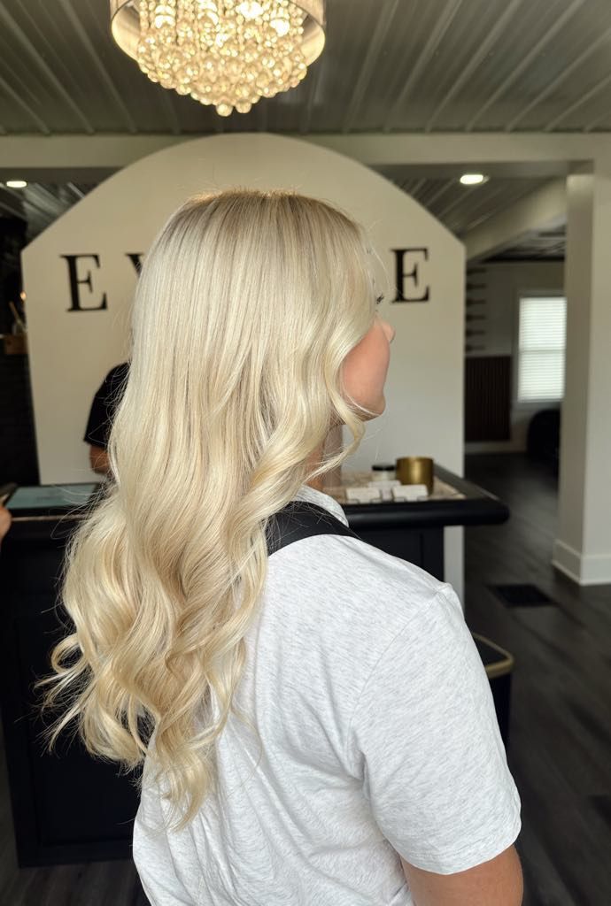 Blonde woman with wavy hair in a salon, facing away from the camera. 