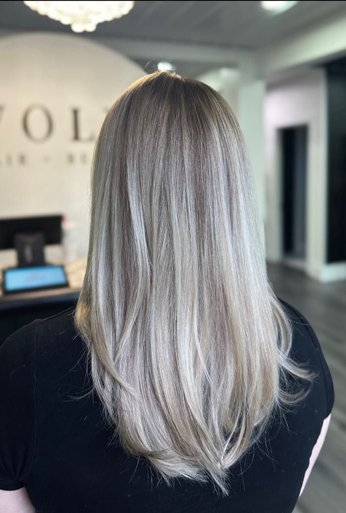 Woman with long, textured ash-blonde hair in a salon setting, facing away from the camera.
