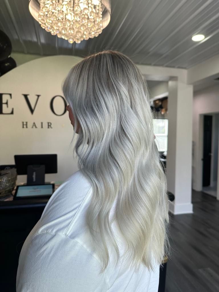 Woman with long, wavy, silver-blonde hair stands in a salon. She wears a white top, and the salon has a black reception desk.