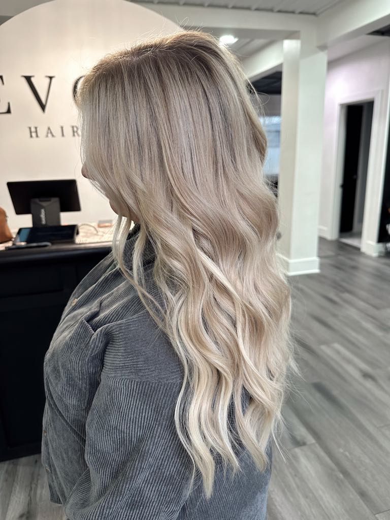 Woman with long wavy blonde hair, standing in a salon. She's wearing a grey textured top.