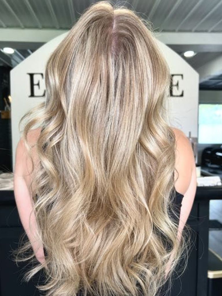 Woman with long, wavy blonde hair stands with her back to the camera in a salon.