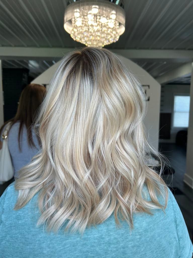 Woman with blonde wavy hair in a salon, wearing a teal shirt, with a bright chandelier overhead.