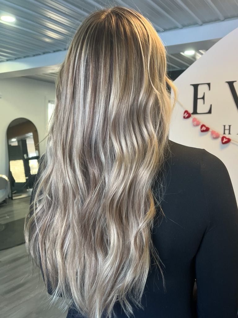Woman with long, wavy, highlighted blonde hair, facing away. Indoors, against a neutral background.