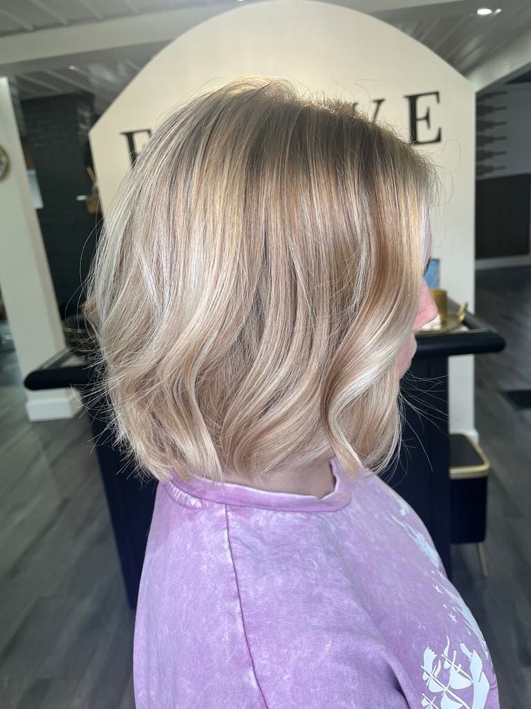 Woman with shoulder-length blonde wavy hair, wearing a light purple shirt, stands inside a salon.