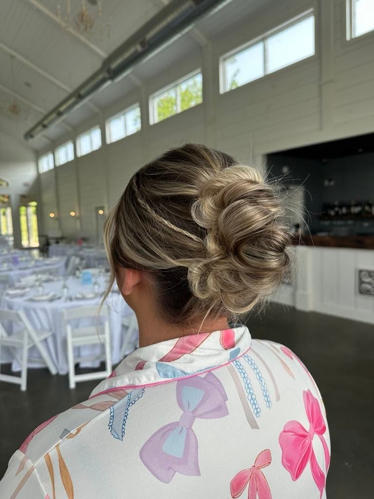 Woman with blonde updo, wearing a bow-print shirt, at an event venue with tables and windows in the background.