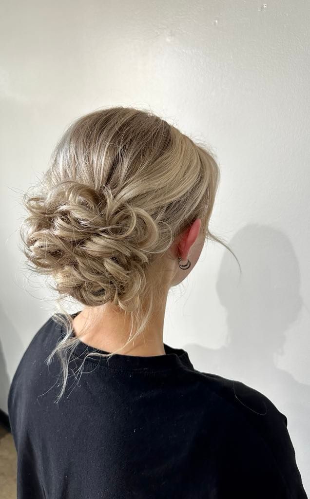 Woman with blonde hair styled in an updo, possibly for a formal occasion, against a white wall.