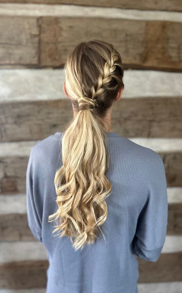 Blonde woman with a braided hairstyle, a side braid and a wavy ponytail. She is wearing a blue sweater, standing in front of a wooden wall.