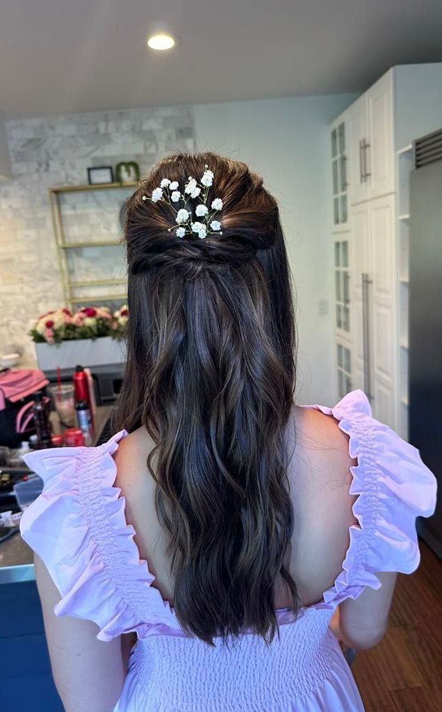 Woman with dark wavy hair, half-up with white flower hairpiece. She wears a lavender dress in a kitchen setting.