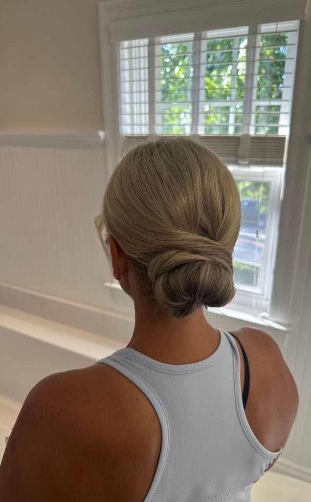 Woman with blonde hair in a low bun, wearing a light blue tank top, standing near a window.