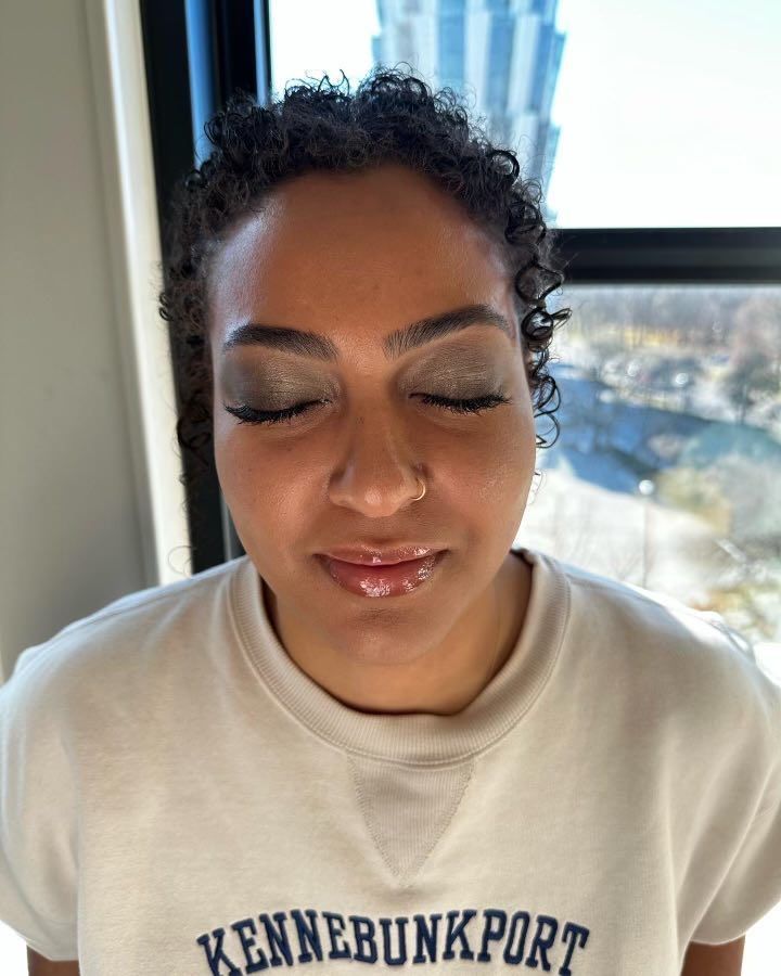 Woman with closed eyes wearing makeup, light sweater, and nose ring, near a window.