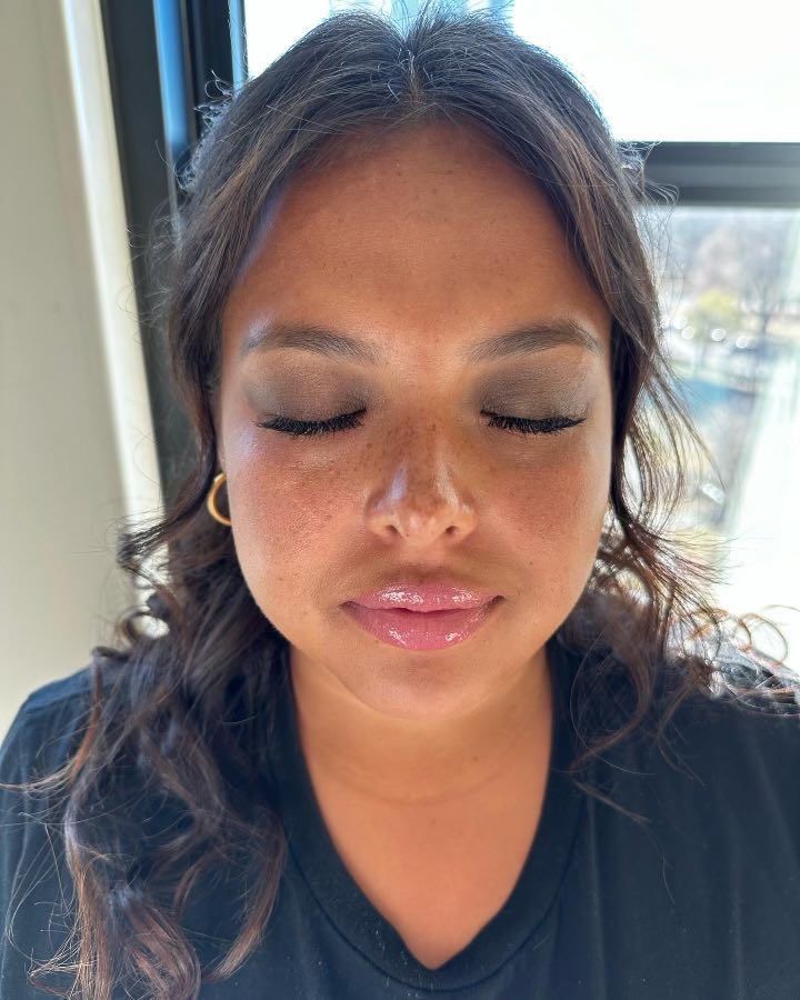 Woman with dark hair and closed eyes, wearing eyeshadow and lip gloss. She is indoors with natural light.