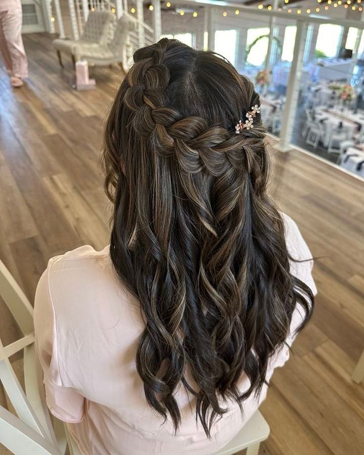Woman with brown hair styled in a half-up braid with loose curls, accented by a small flower hair clip, seated indoors.