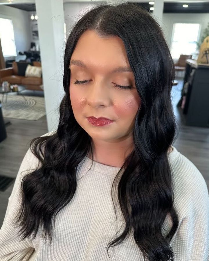 Woman with dark wavy hair and makeup, eyes closed, wearing a cream-colored sweater. Indoors, possibly a salon.