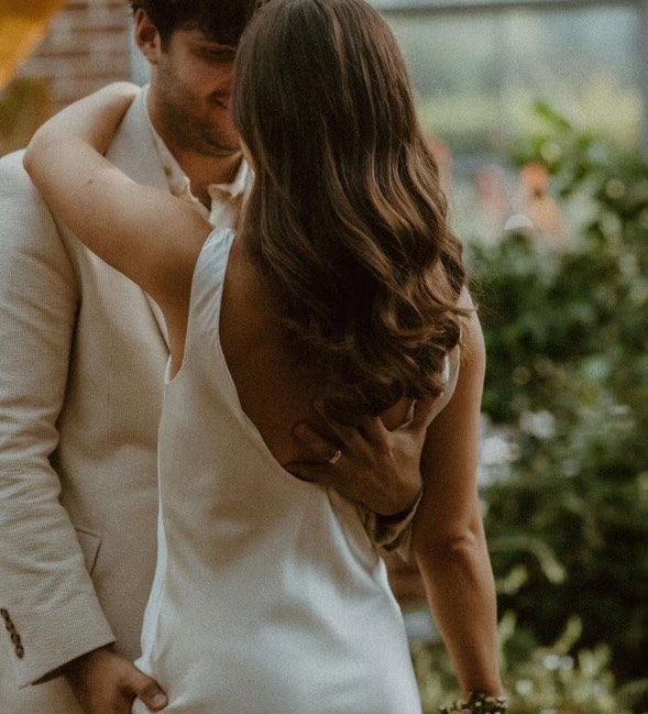 Woman in white dress embraced by a man in a tan suit, likely outdoors, with greenery in the background.