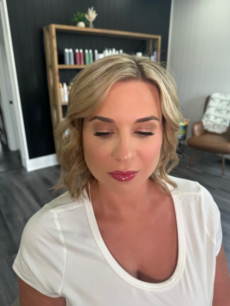 Woman with blonde wavy hair and professional makeup, wearing a white shirt, in a salon setting.
