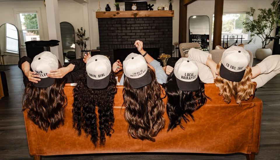 Five people wearing baseball caps lie on a brown couch. Long, wavy brown hair cascades down.