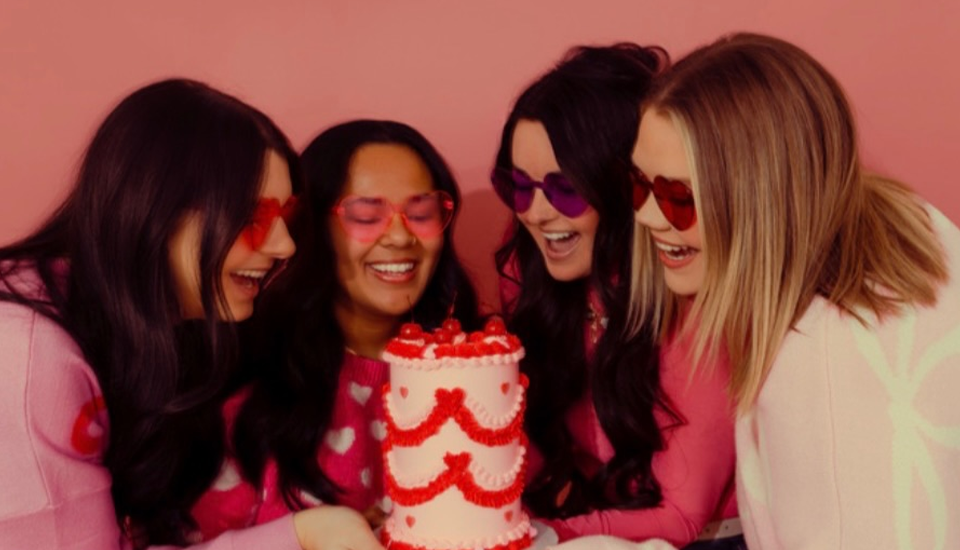 Four women in pink look excitedly at a heart-decorated cake. Pink background.