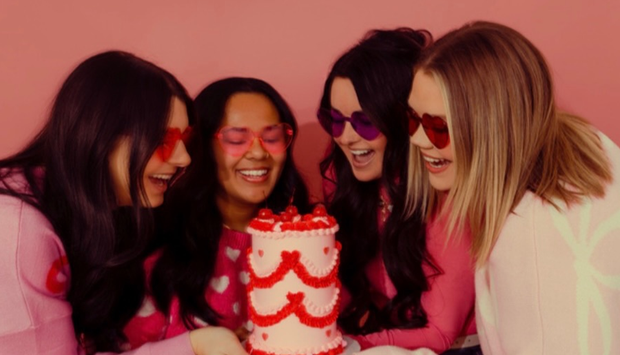 Four women in pink look excitedly at a heart-decorated cake. Pink background.