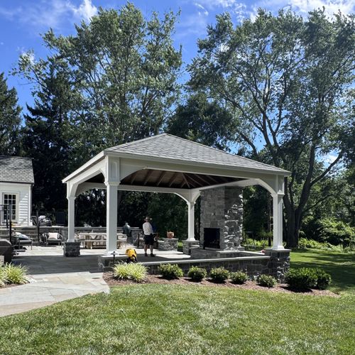 Outdoor pavilion with stone columns, people seated inside, and a landscaped lawn under sunny trees