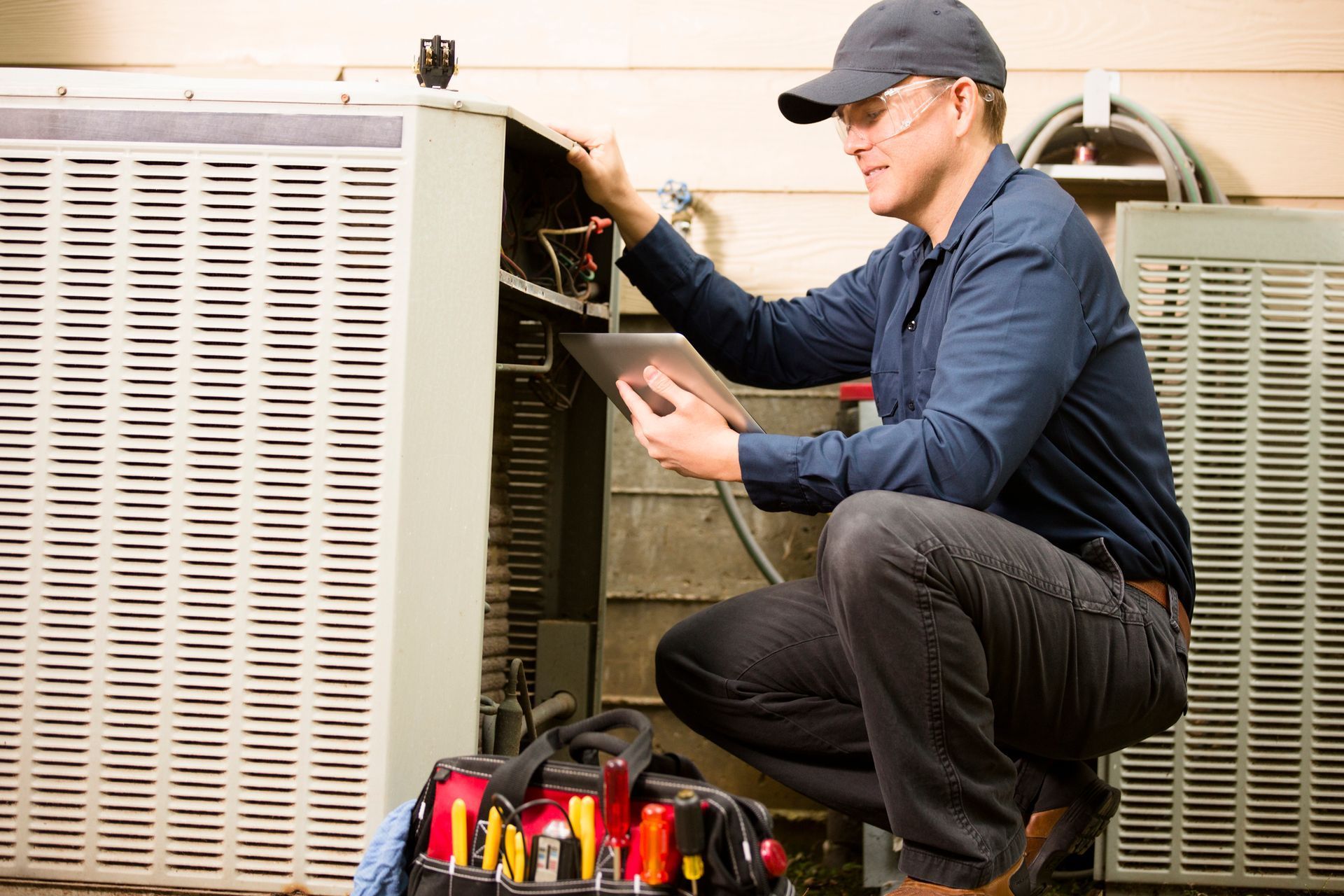 HVAC technician in cap squats near an air conditioning unit, using a tablet, with tools.