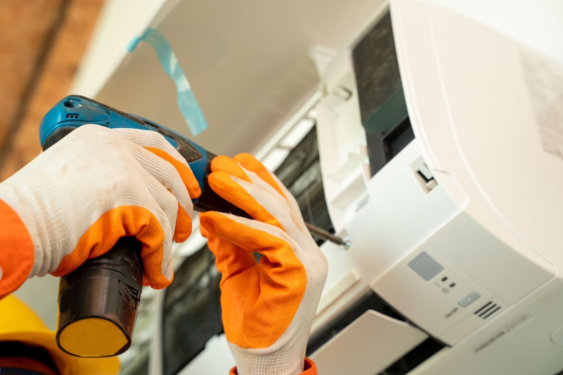Hands in orange work gloves, using a drill to service a white air conditioner unit.