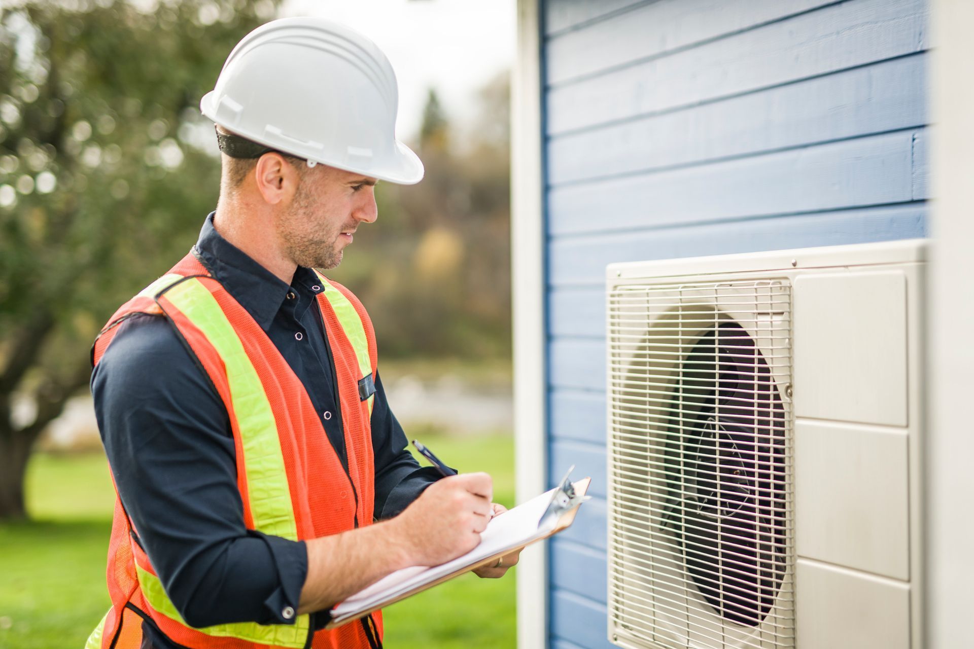 HVAC technician in hard hat and vest inspecting an outdoor air conditioning unit, writing on a clipboard.