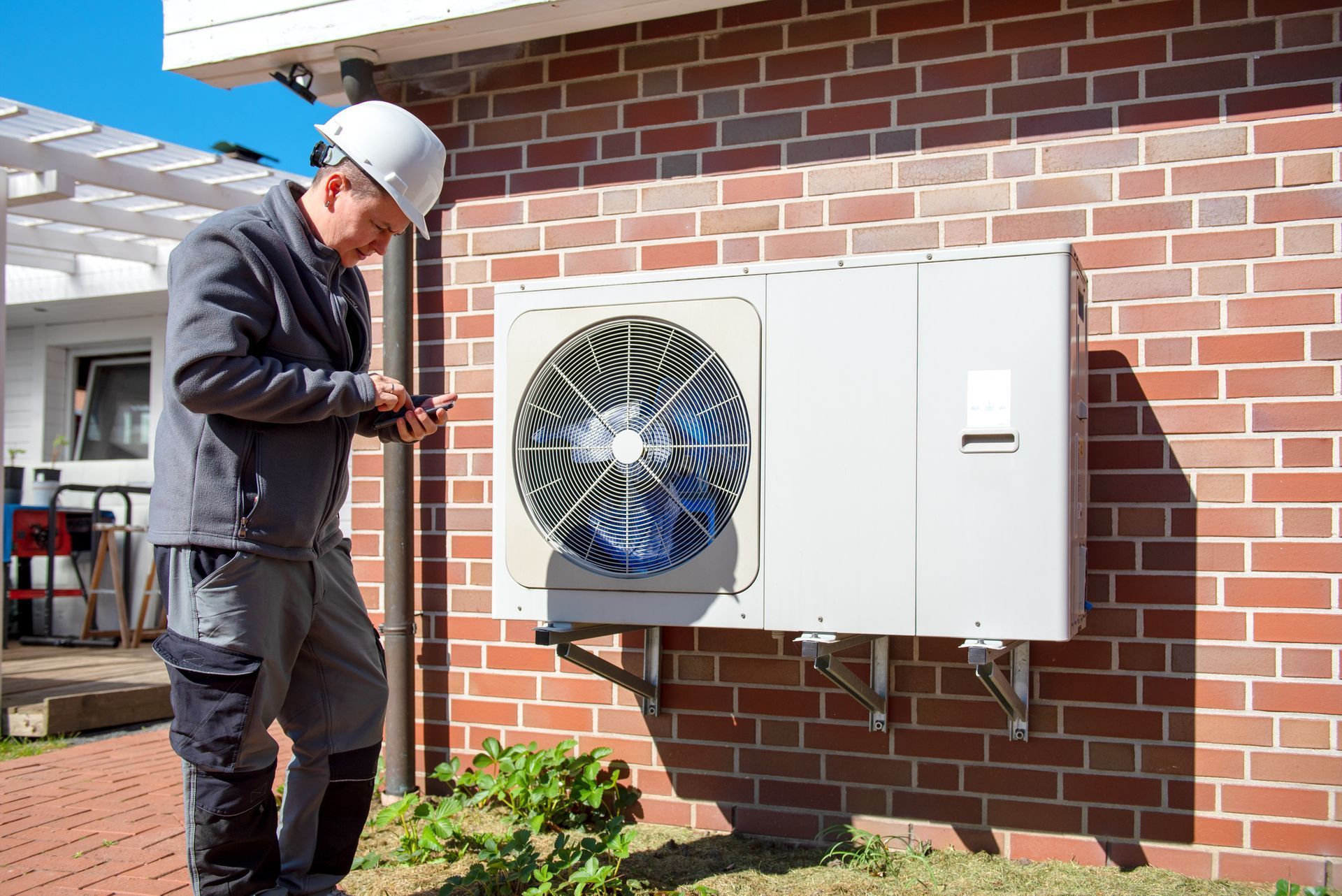 Technician in hard hat inspecting an HVAC unit mounted on a brick wall.
