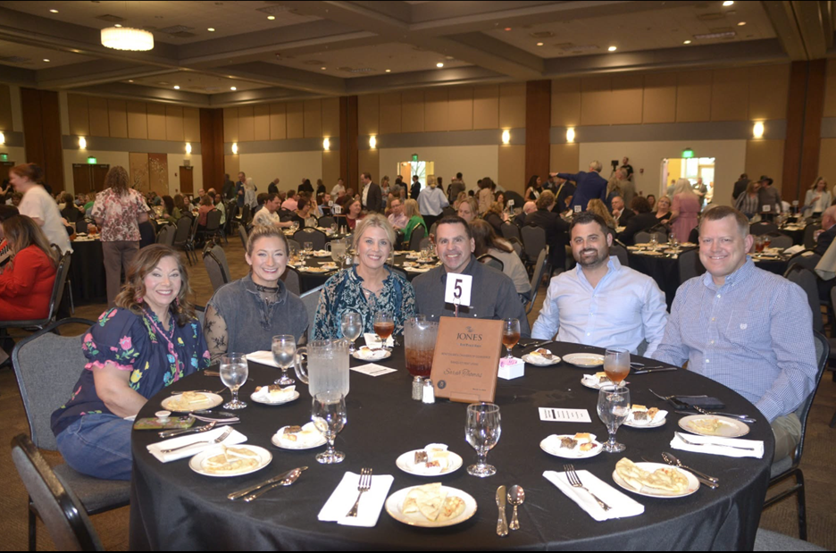 A group of six people sit around a round dinner table in a large banquet hall, smiling for a photo at a formal event.