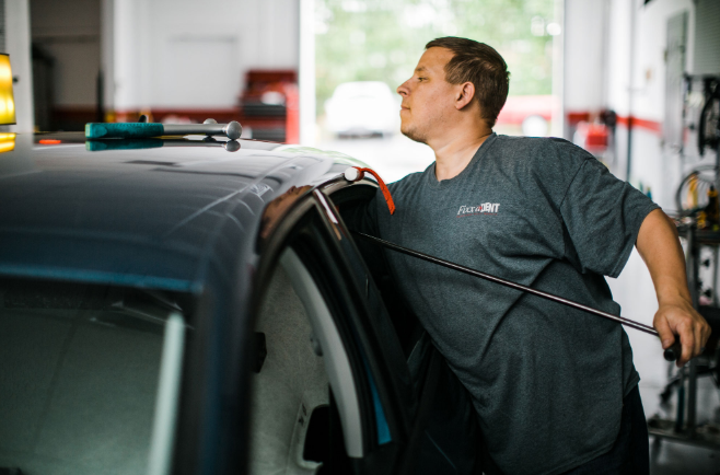 Mechanic working on a car in a garage, using a tool to access inside, wearing a gray shirt.