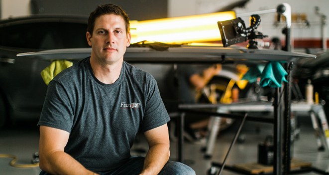 Man in gray shirt in auto body shop. He sits near a car and work table.