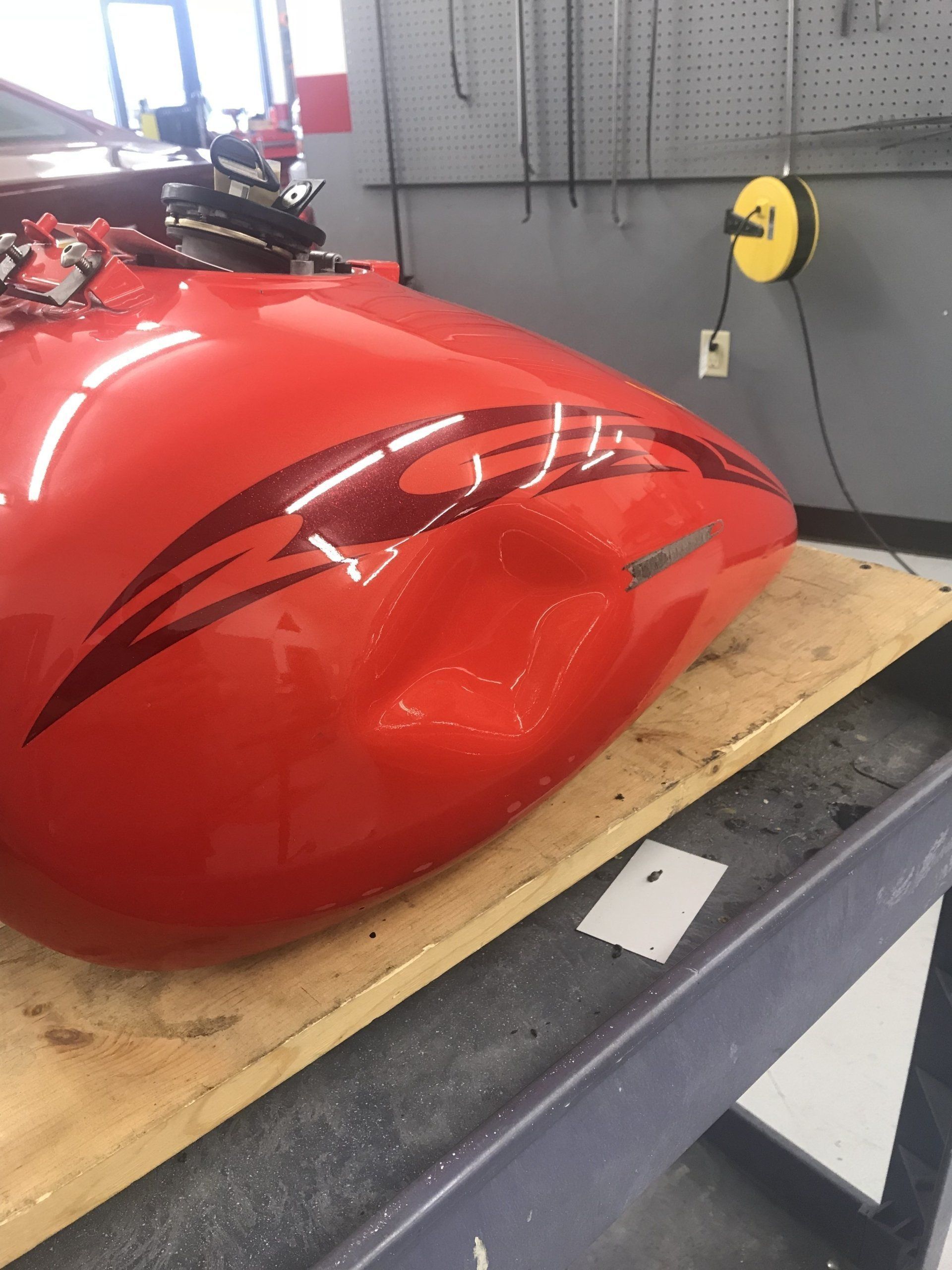 Red motorcycle gas tank with a dent, resting on a wooden surface in a garage.