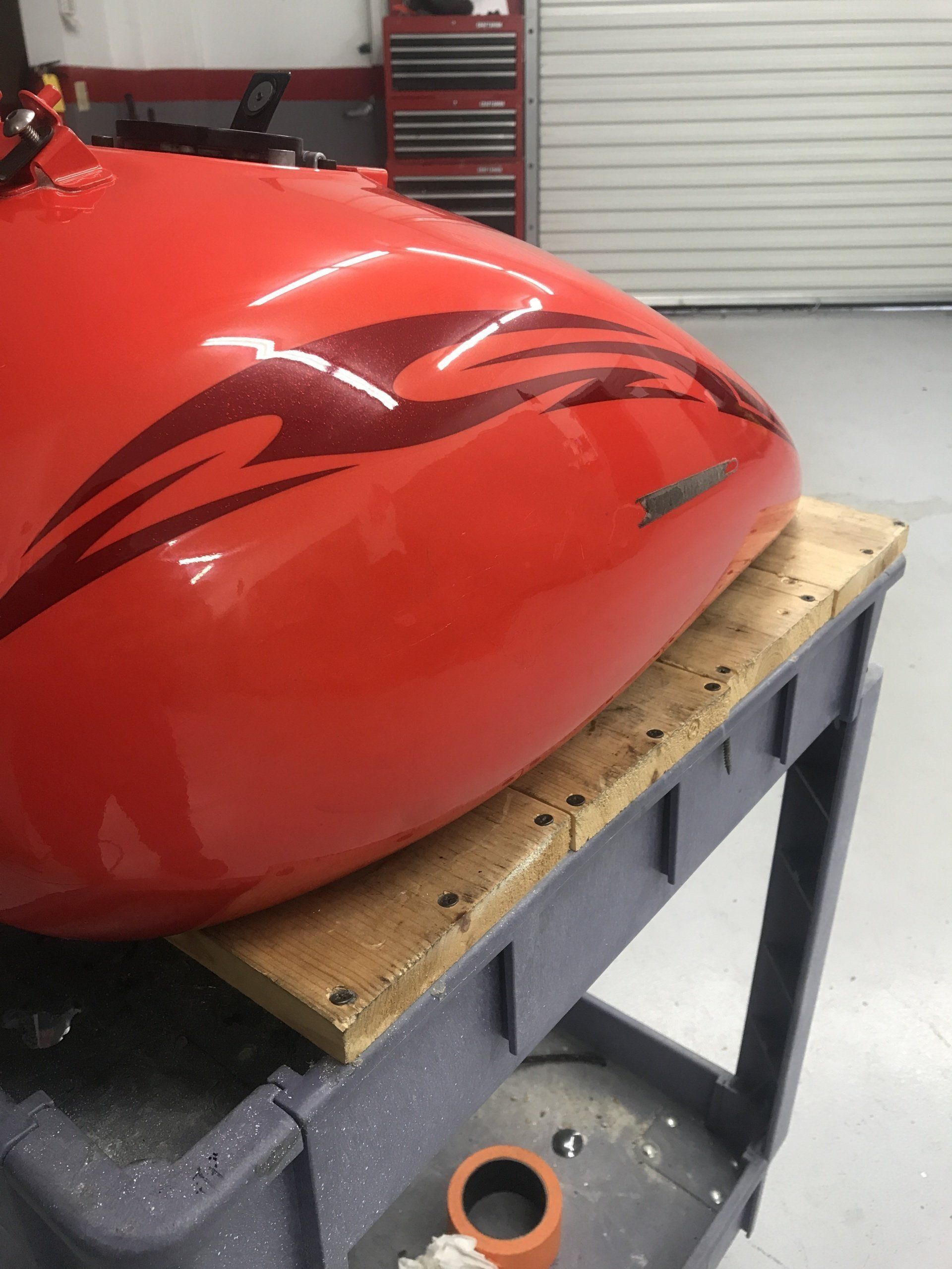 Red motorcycle gas tank with dark tribal design on a wooden surface over a grey cart in a shop.