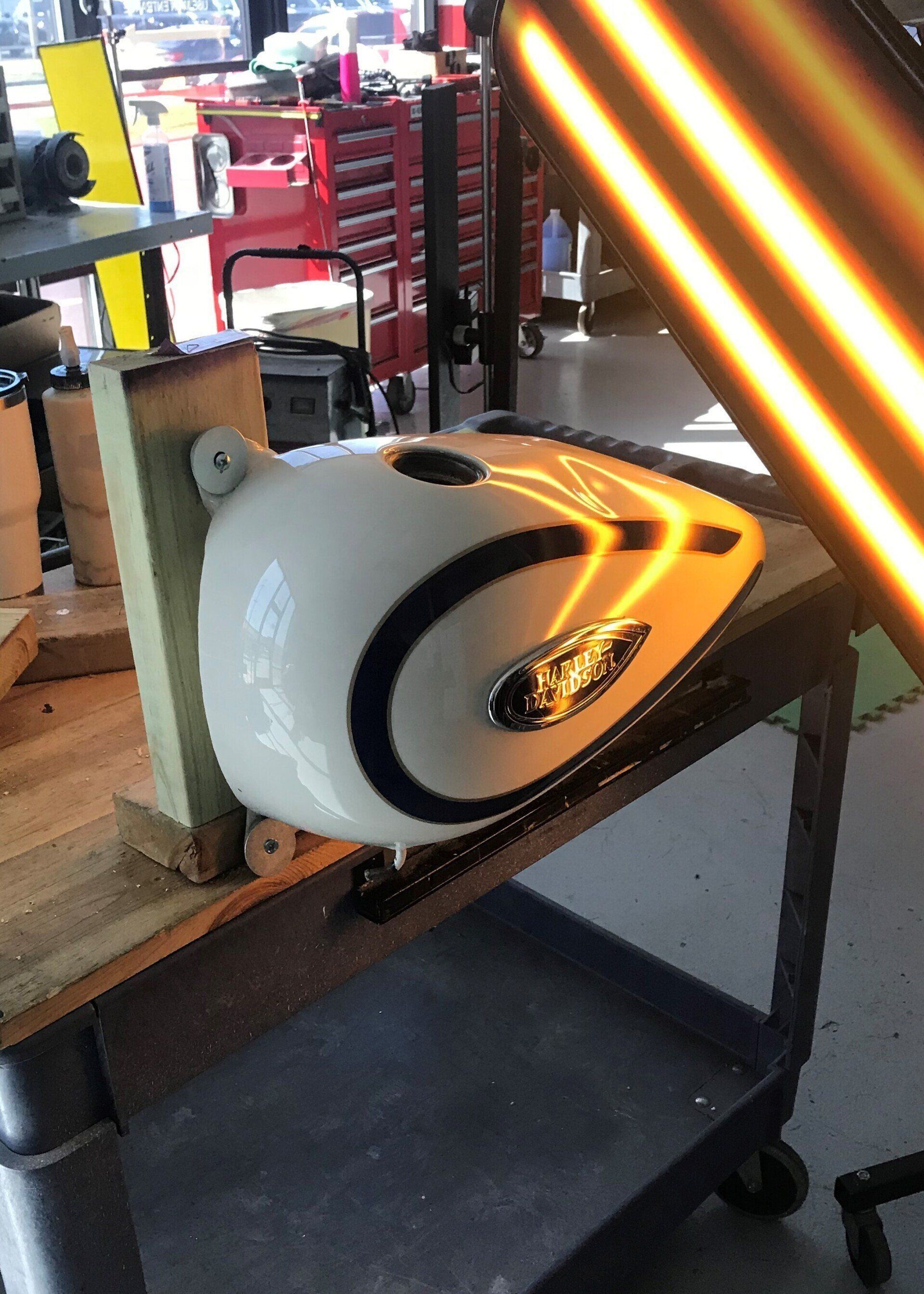 Cream-colored motorcycle gas tank on a cart in a workshop; a light source highlights its curve.