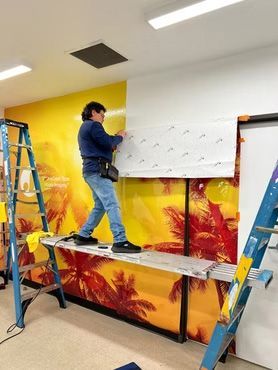 Worker installing wall art on a large wall beside ladders in a bright room