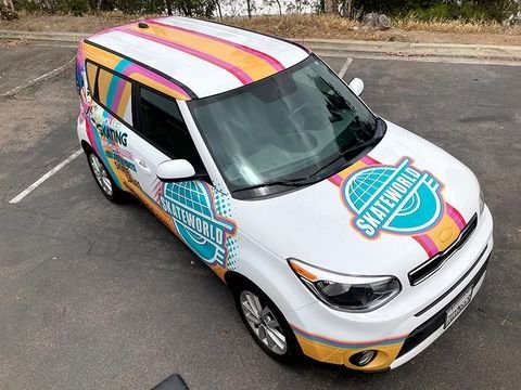 A colorfully wrapped white compact car is parked on the pavement, viewed from above.