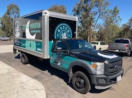 A teal and black delivery truck with a logo parked outdoors in a sunny lot