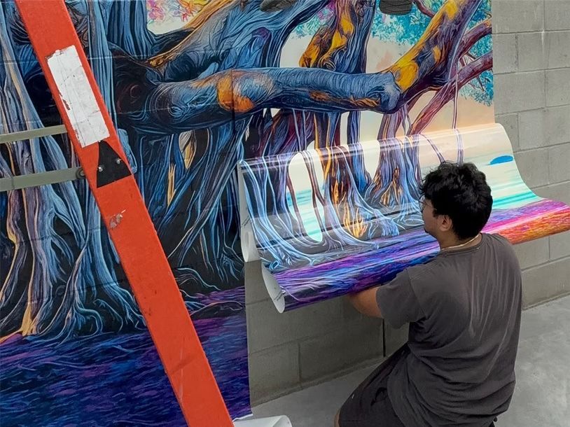 An artist wrapping a colorful mural around a wall, using a ladder and pasted print panels