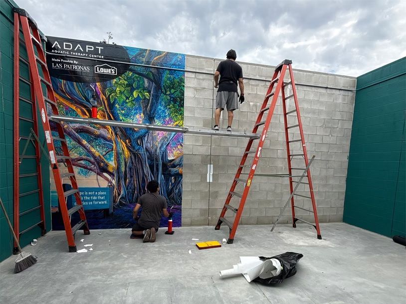 People painting a colorful mural on a concrete wall, with ladders and supplies in a courtyard.