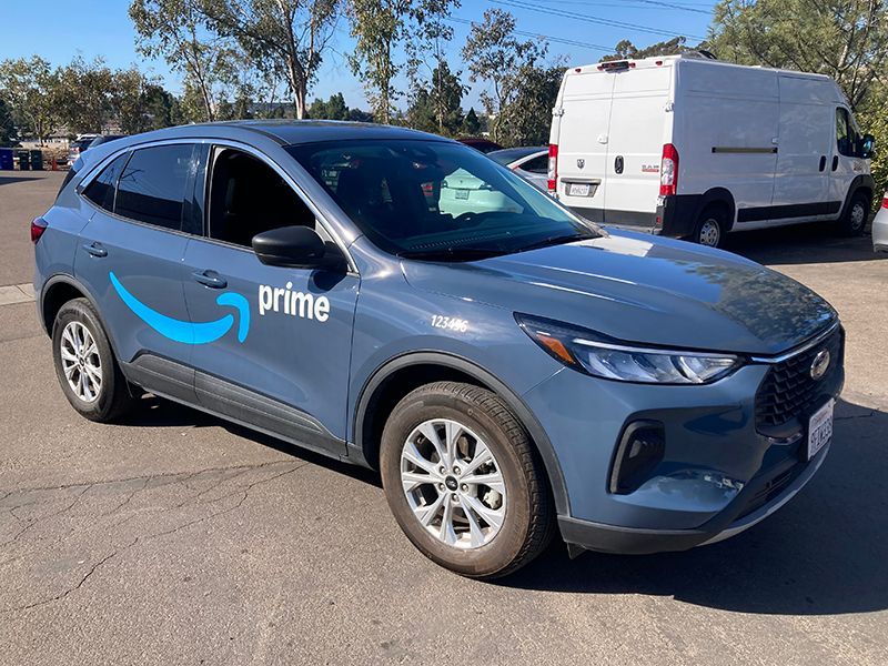 Blue Amazon Prime delivery van parked outdoors on a sunny street, with a white van in the background