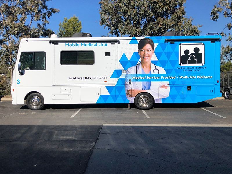 Blue and white medical outreach bus parked outdoors with health messaging and portraits on the side.