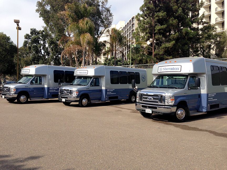 Three blue and white shuttle vans parked in a lot near trees and apartment buildings