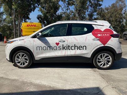 White mama's kitchen delivery car with pink heart logo parked outdoors near a DHL van.