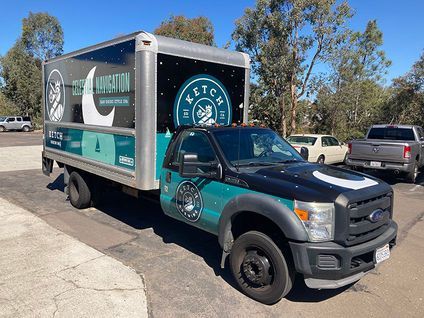 Teal-and-black moving truck with a large branded box parked on a sunny street
