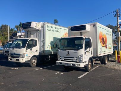 Two white delivery trucks parked side by side outdoors, one with fruit graphics on the trailer.