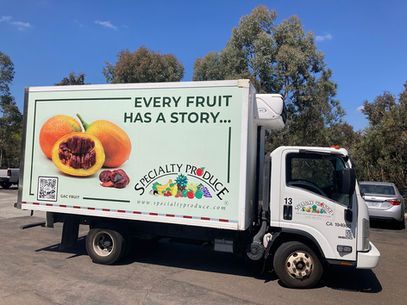 A produce truck with a fruit graphic and the Every fruit has a story slogan on a sunny street.