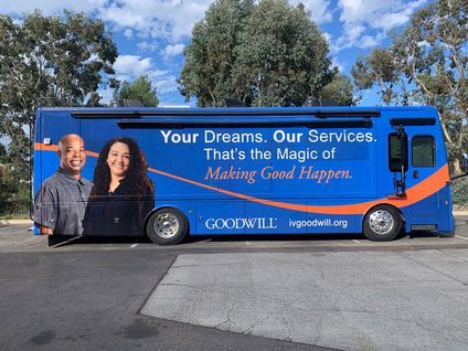 Blue Goodwill bus with a charity slogan and two smiling people on the side, parked outdoors under trees.